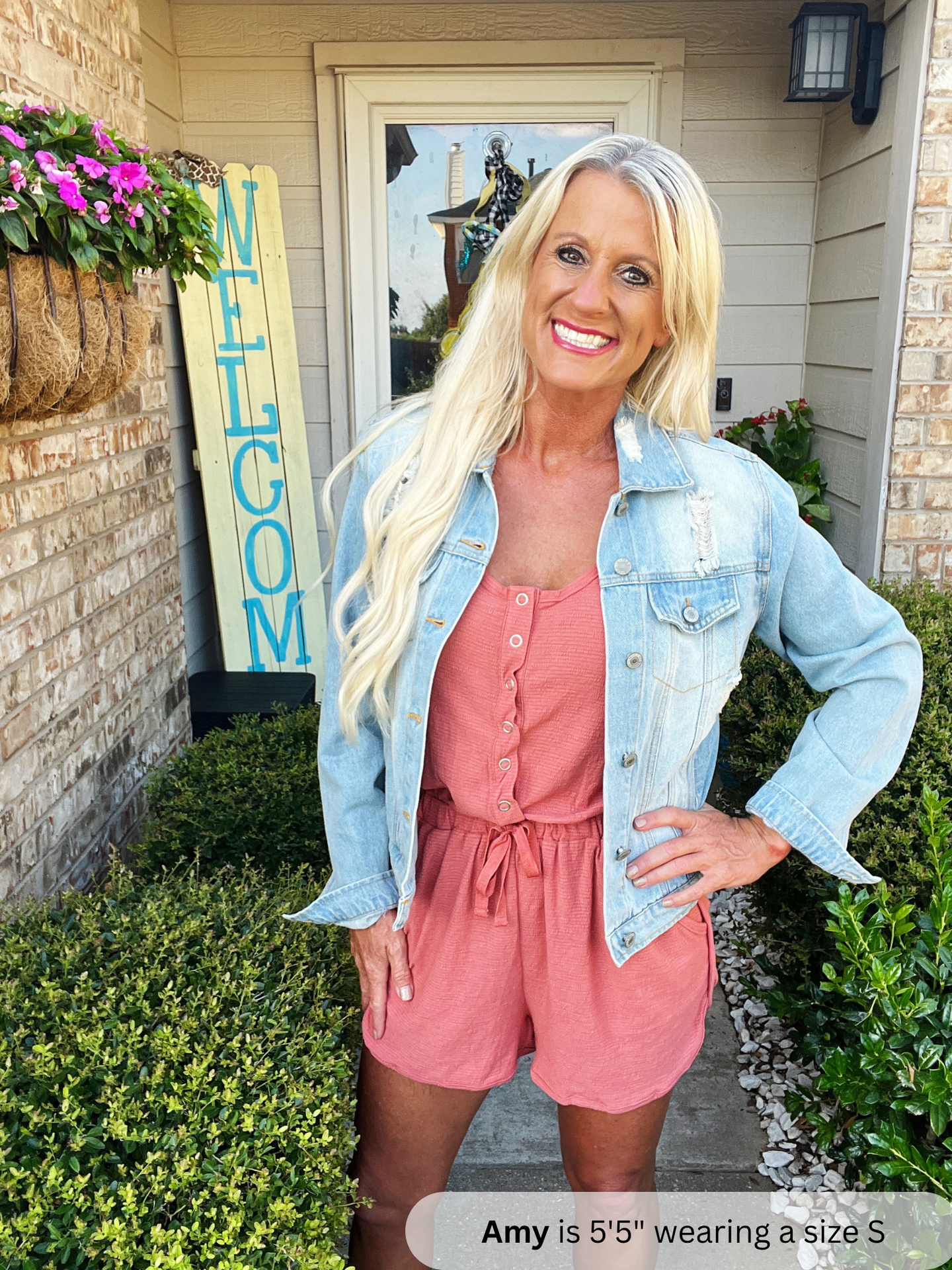 /Rust/ Woman in pink romper and denim jacket standing in front of a 'Welcome' sign.