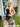 Woman in black tank top and leather shorts standing on a porch with plants and a door in the background.