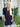 Woman wearing a black dress standing outside a house with plants and a 'WELCOME' sign.