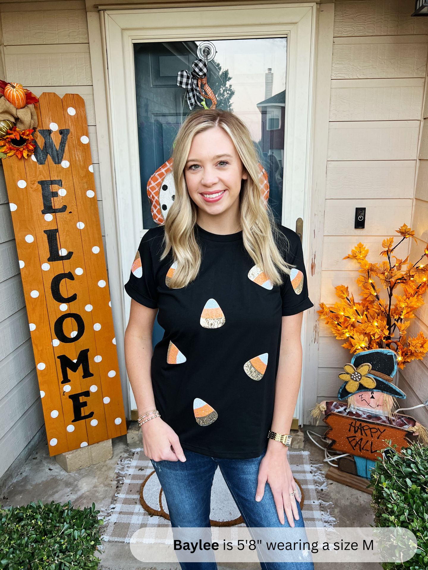 Woman wearing a black t-shirt with candy corn patterns in front of a house decorated for Halloween.