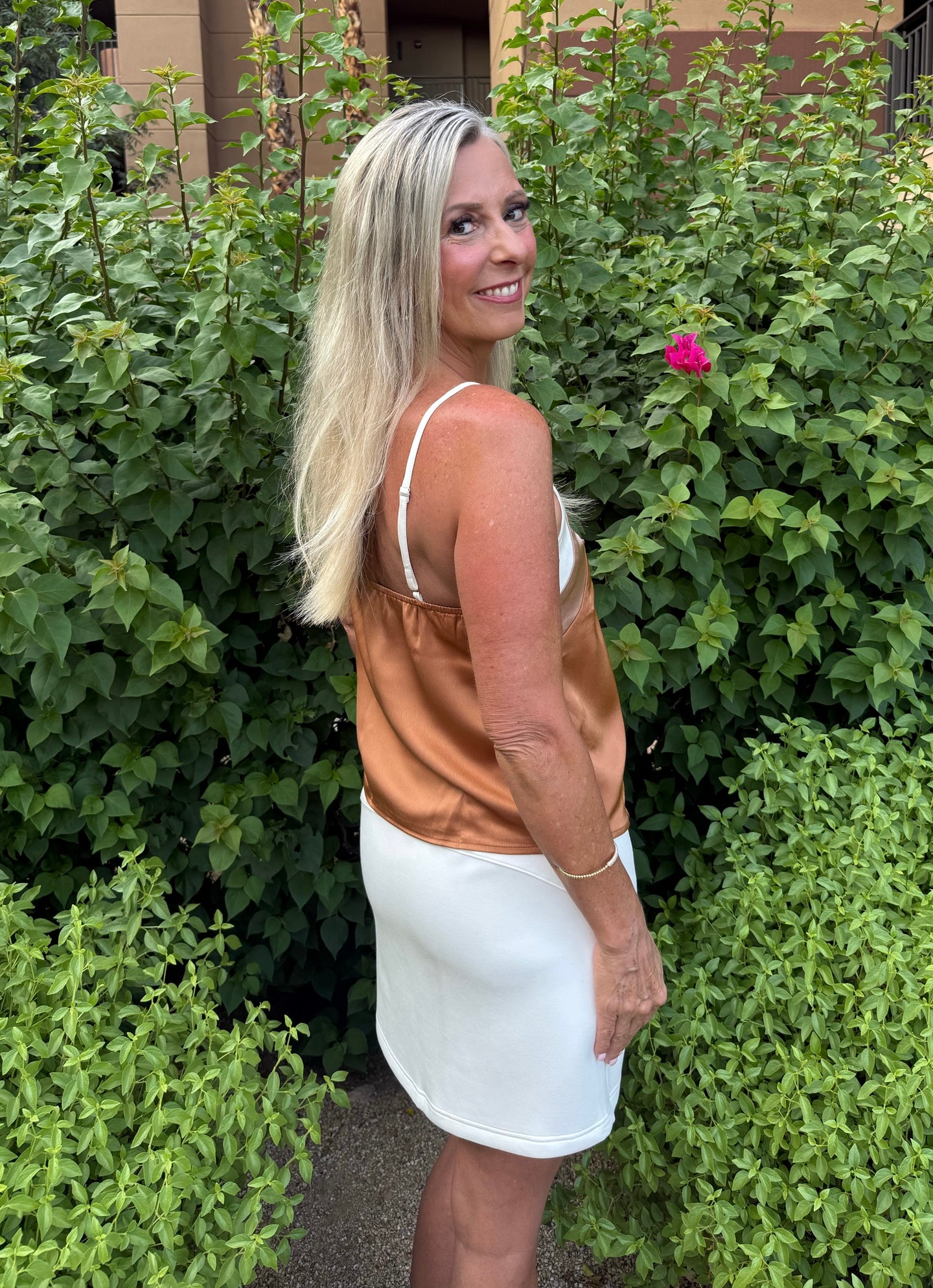 Woman in a white dress standing in front of green bushes with a pink flower.