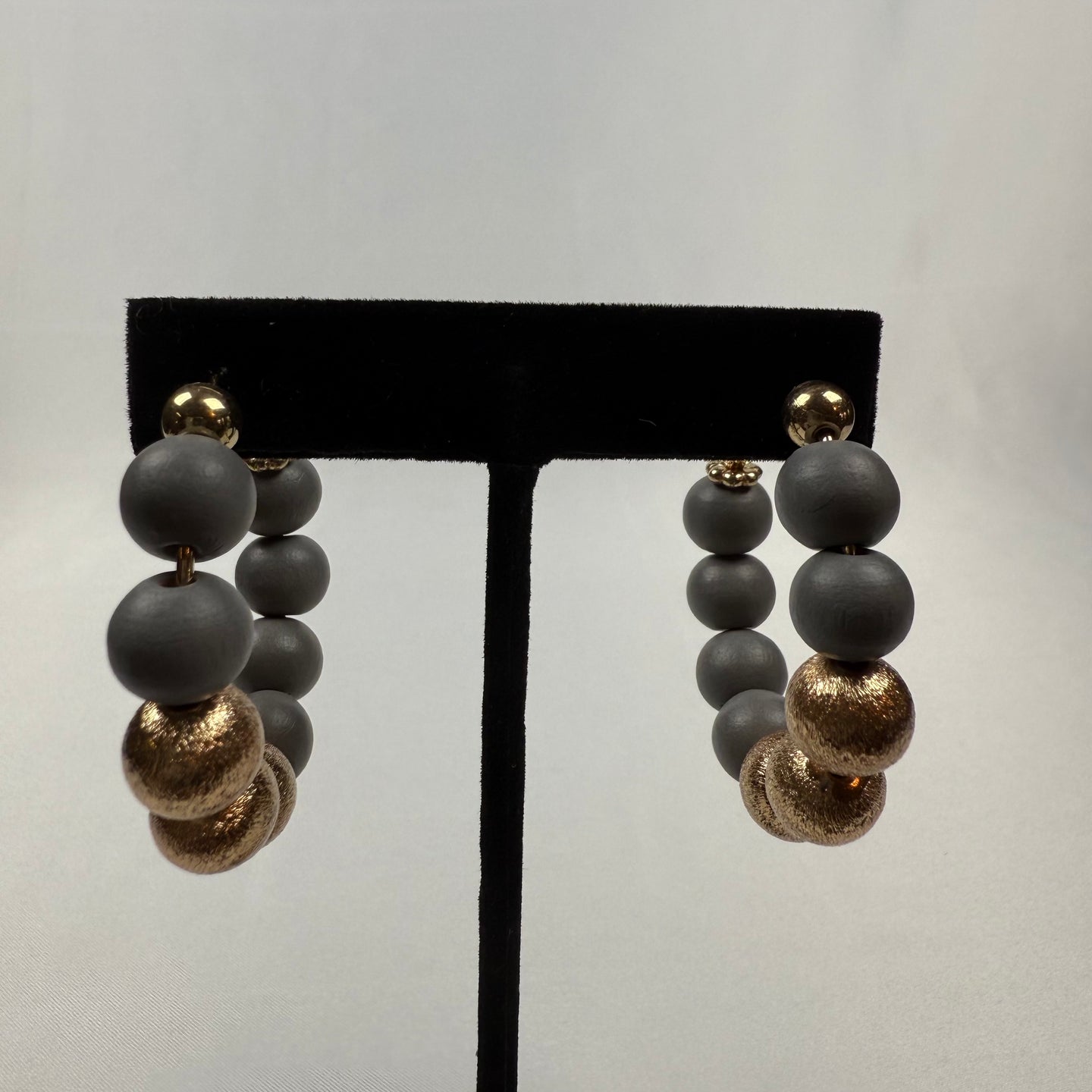 A pair of hoop earrings made of matte gray beads and textured gold beads, displayed on a black stand against a plain, light gray background.