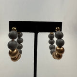A pair of hoop earrings made of matte gray beads and textured gold beads, displayed on a black stand against a plain, light gray background.