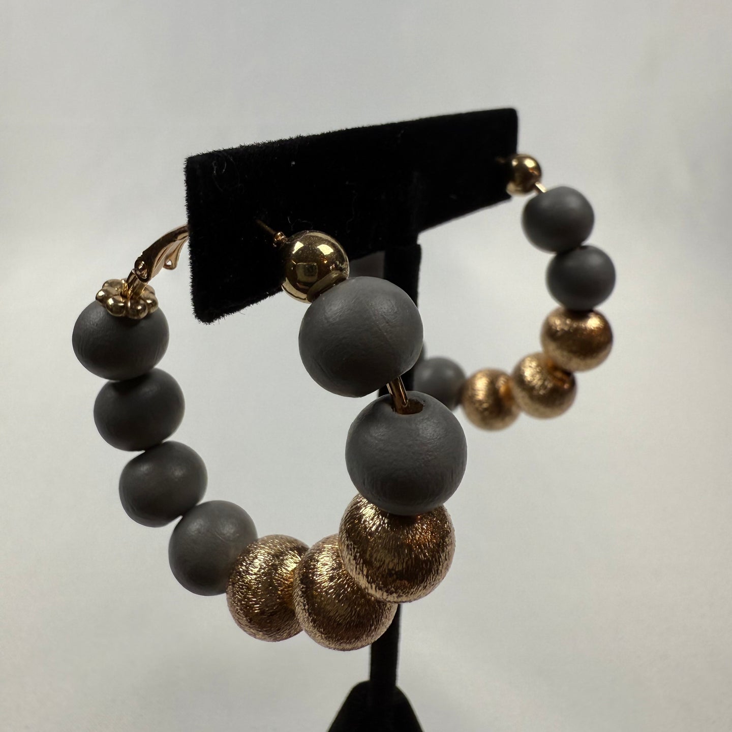A pair of hoop earrings displayed on a black stand, featuring alternating large matte gray beads and textured metallic gold beads, with gold accents at the clasp.