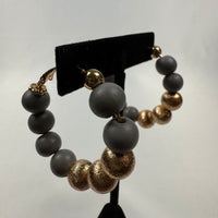 A pair of hoop earrings displayed on a black stand, featuring alternating large matte gray beads and textured metallic gold beads, with gold accents at the clasp.
