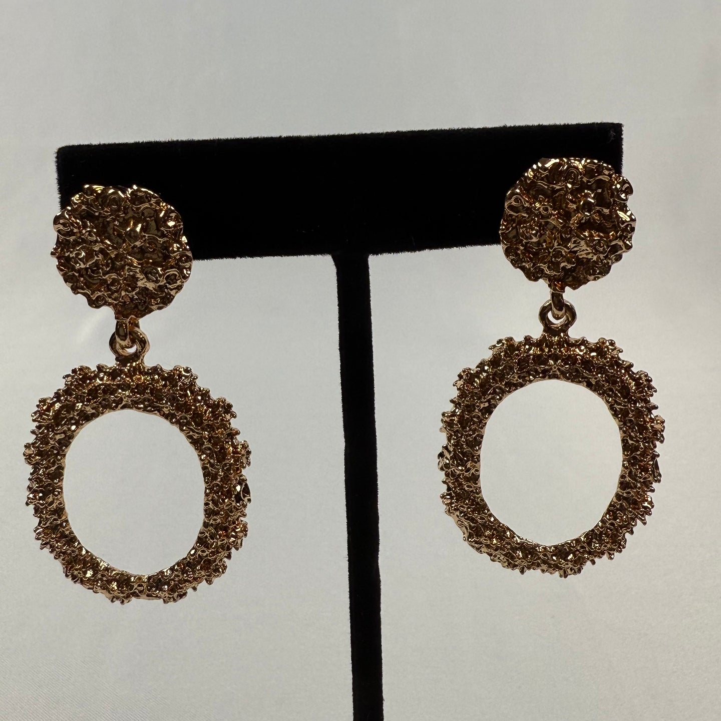 Gold textured dangle earrings with circular studs and larger open oval pendants, both featuring a rough, organic surface, displayed on a black stand against a plain light background.
