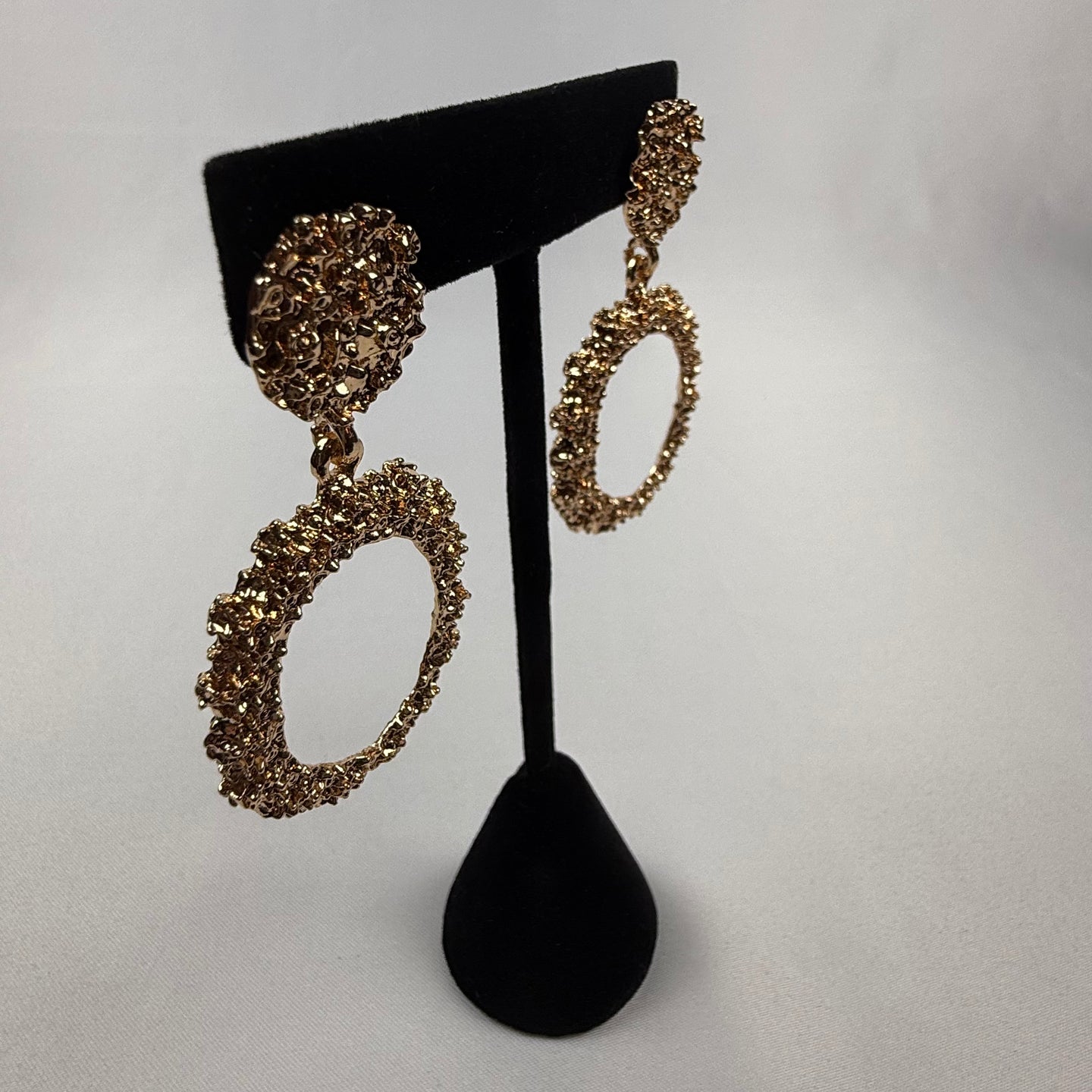 Gold textured hoop earrings on a black velvet stand, displayed against a plain white background. The earrings feature a circular, intricate design with a rough, sparkling finish.