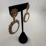Gold textured hoop earrings on a black velvet stand, displayed against a plain white background. The earrings feature a circular, intricate design with a rough, sparkling finish.