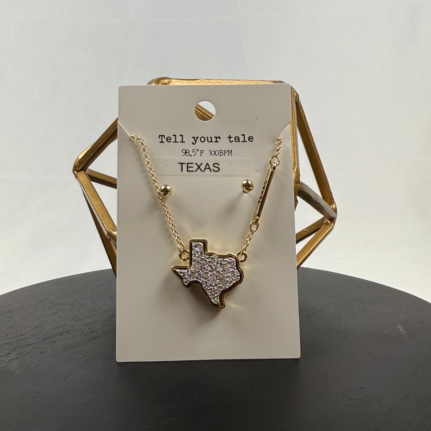 A gold necklace with a Texas-shaped pendant covered in rhinestones is displayed on a card that reads Tell your tale, 98.5° F 100 BPM, TEXAS. The display sits on a black surface with a gold geometric decor piece behind it.