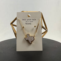 A gold necklace with a Texas-shaped pendant covered in rhinestones is displayed on a card that reads Tell your tale, 98.5° F 100 BPM, TEXAS. The display sits on a black surface with a gold geometric decor piece behind it.