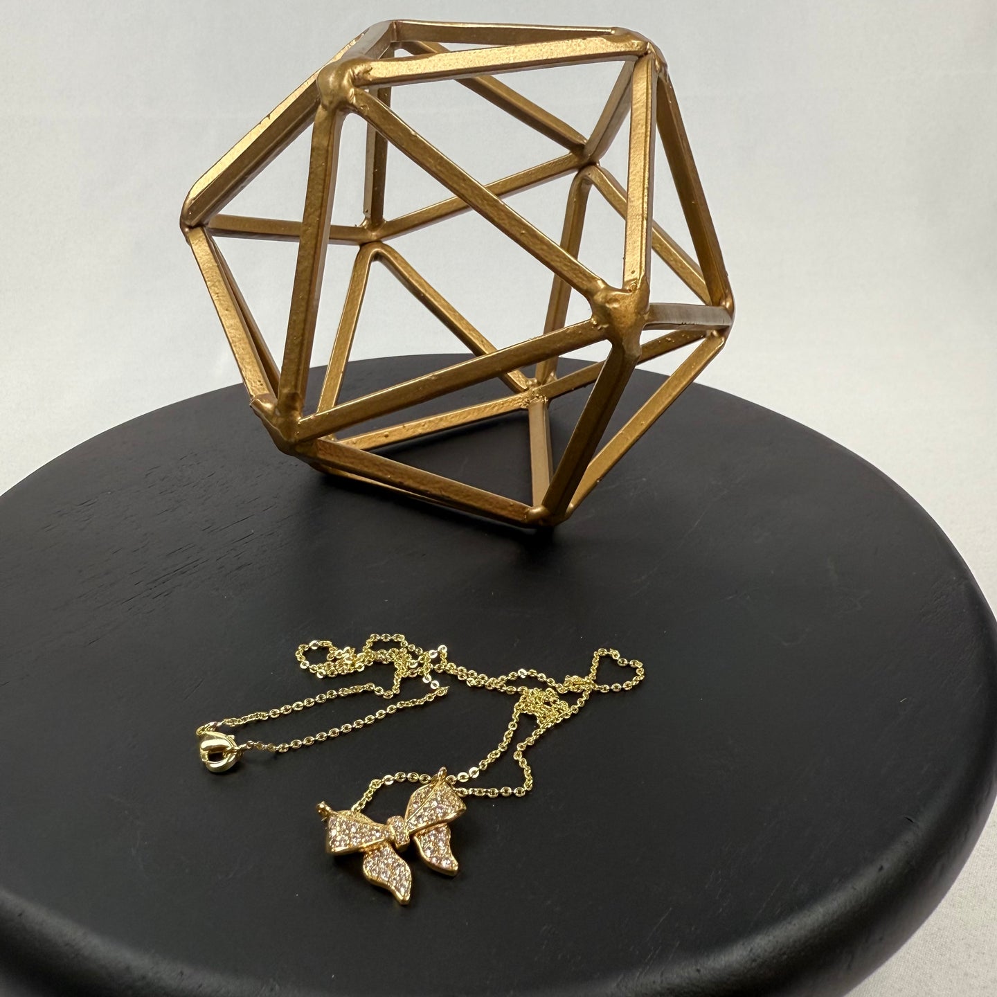A gold geometric sculpture sits on a black round surface next to a delicate gold necklace with a butterfly pendant adorned with small crystals.