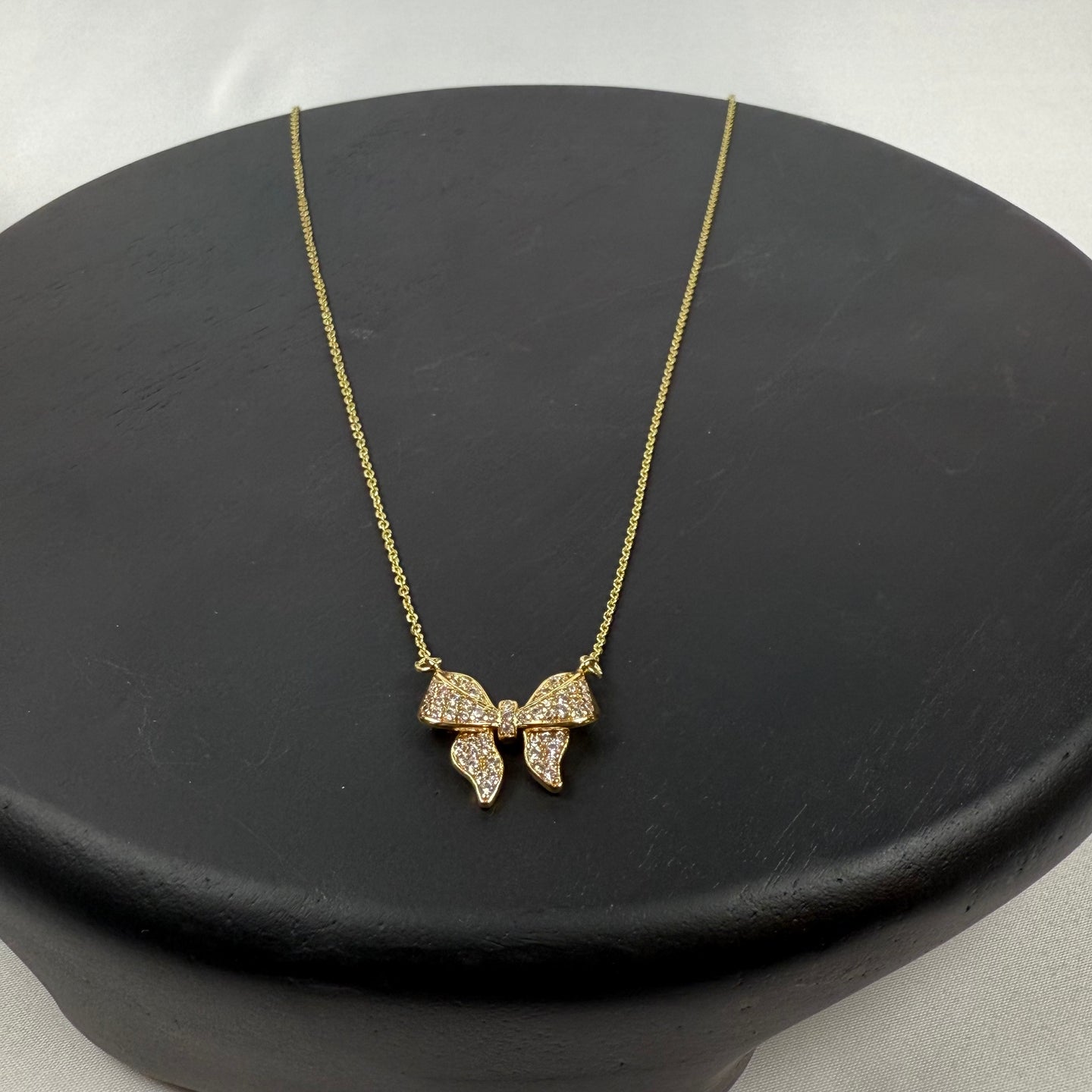 A delicate gold necklace with a butterfly-shaped pendant adorned with small sparkling crystals, displayed on a matte black circular stand against a light background.
