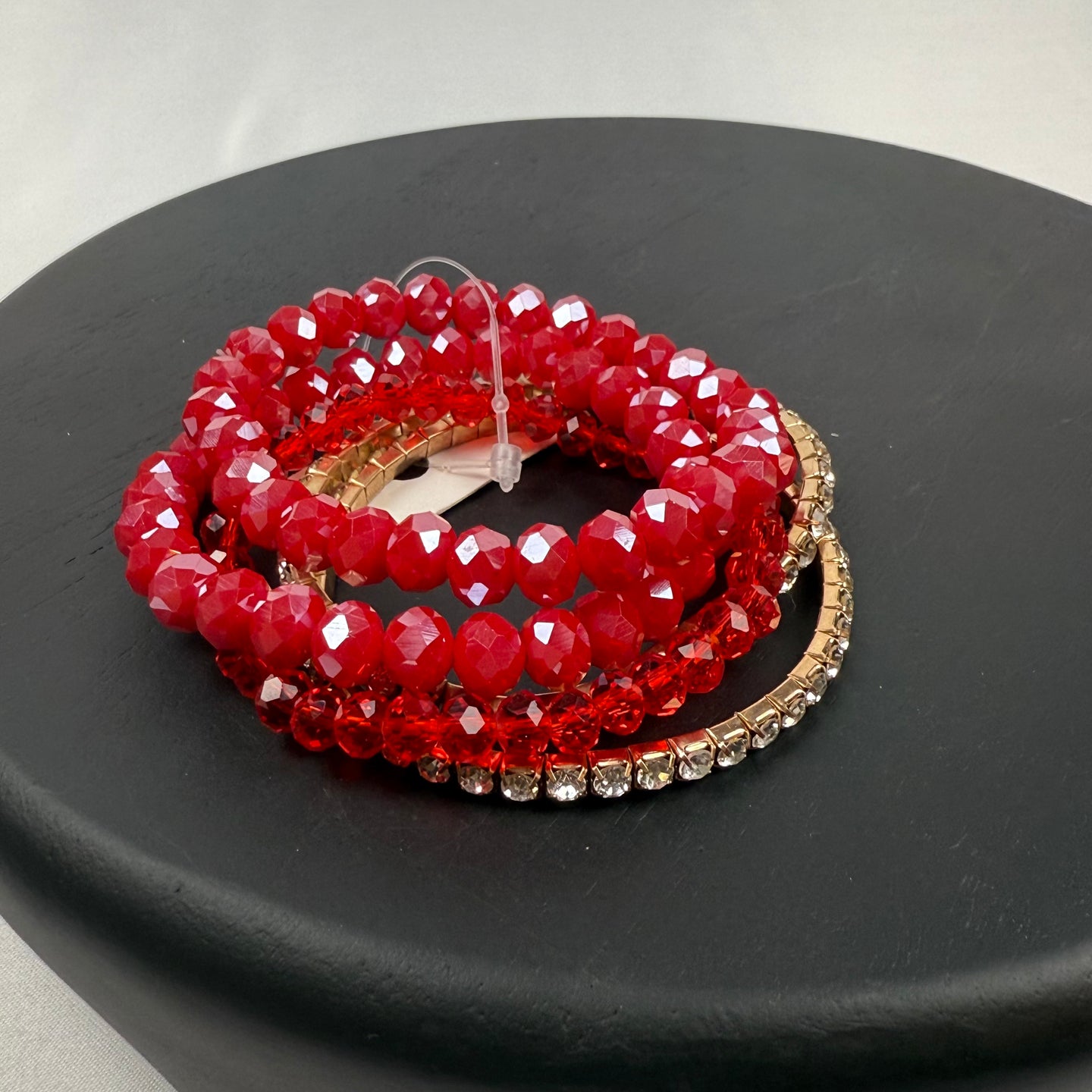 A stack of red faceted beaded bracelets and a gold bracelet with clear rhinestones, displayed on a round black surface against a light background.