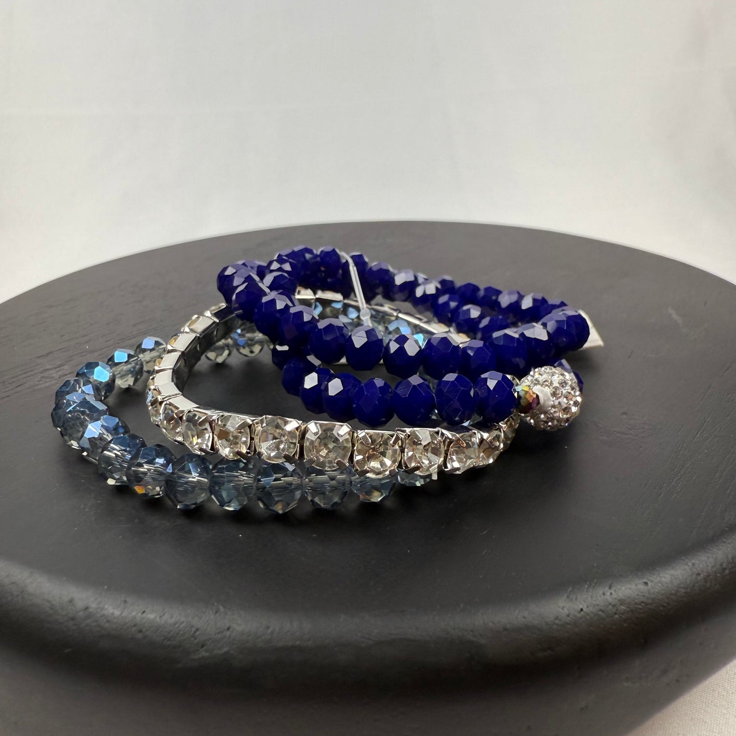 Three beaded bracelets—one blue, one clear, and one dark blue with a crystal bead—are stacked on a round black platform against a plain white background.