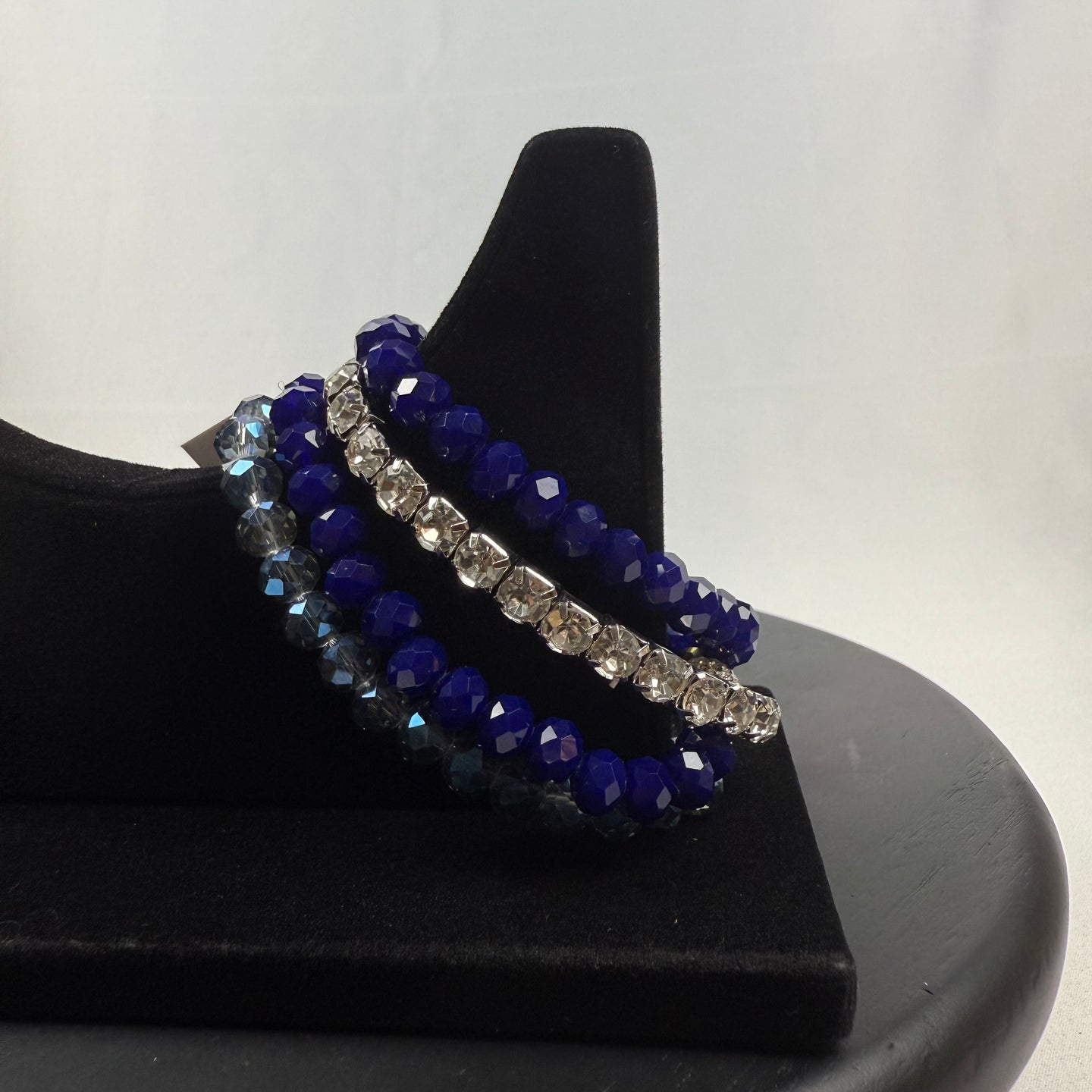 A black velvet bracelet display holds three beaded bracelets: one with clear faceted beads, one with dark blue faceted beads, and one with alternating blue and silver beads, against a plain white background.