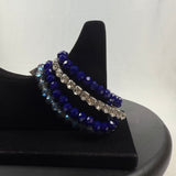 A black velvet bracelet display holds three beaded bracelets: one with clear faceted beads, one with dark blue faceted beads, and one with alternating blue and silver beads, against a plain white background.