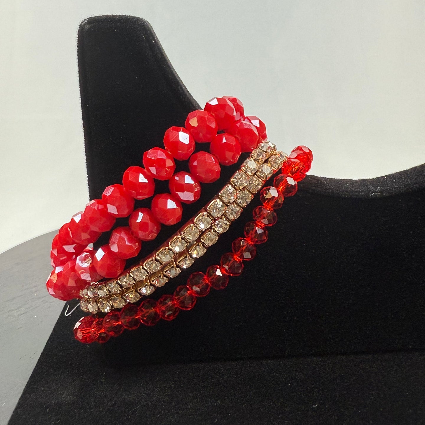A set of four beaded bracelets displayed on a black stand: two with large faceted red beads, one with small clear rhinestones, and one with small red beads.