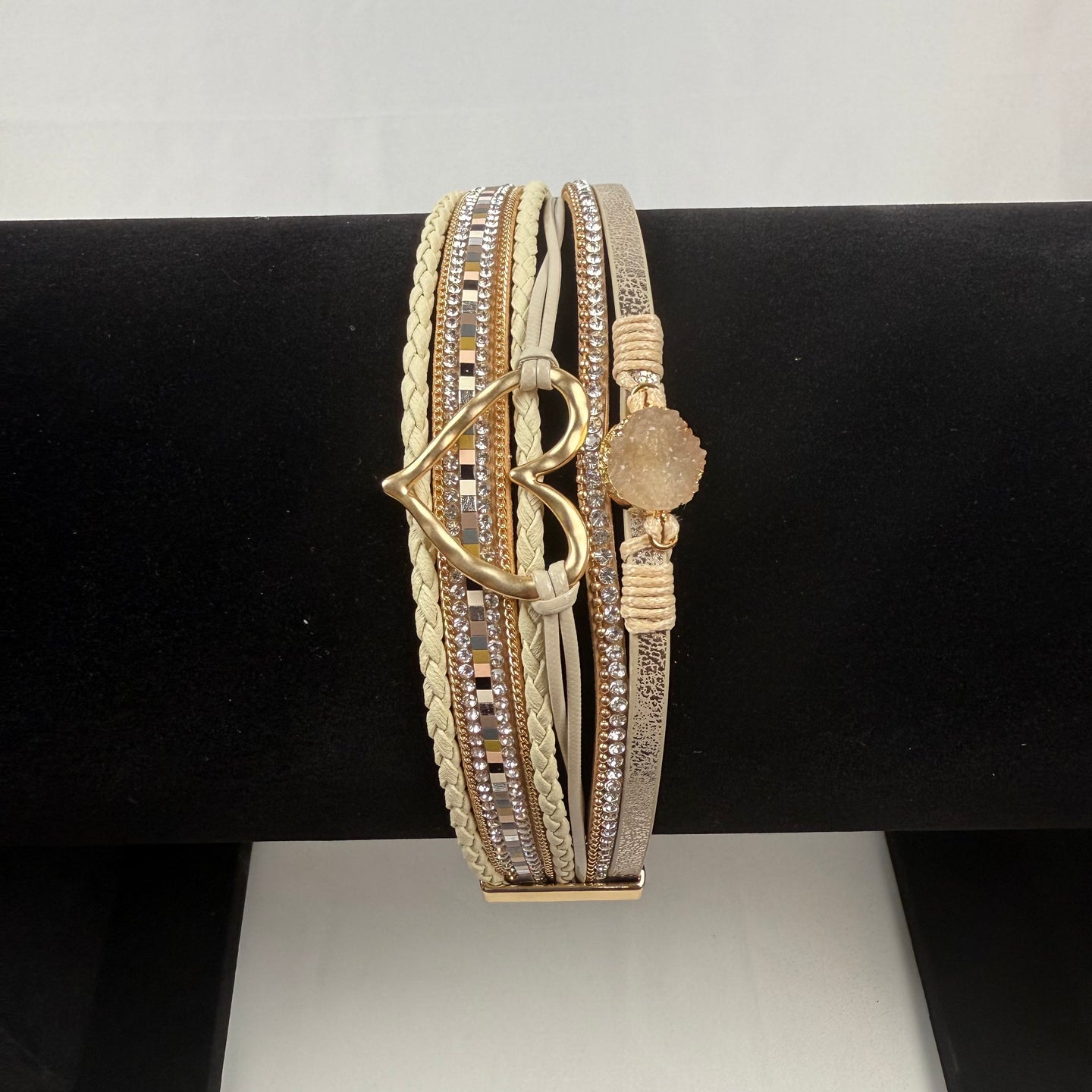 A multi-strand bracelet featuring gold, silver, and white bands with various textures, crystal accents, a heart-shaped charm, and a round stone on a black display stand.