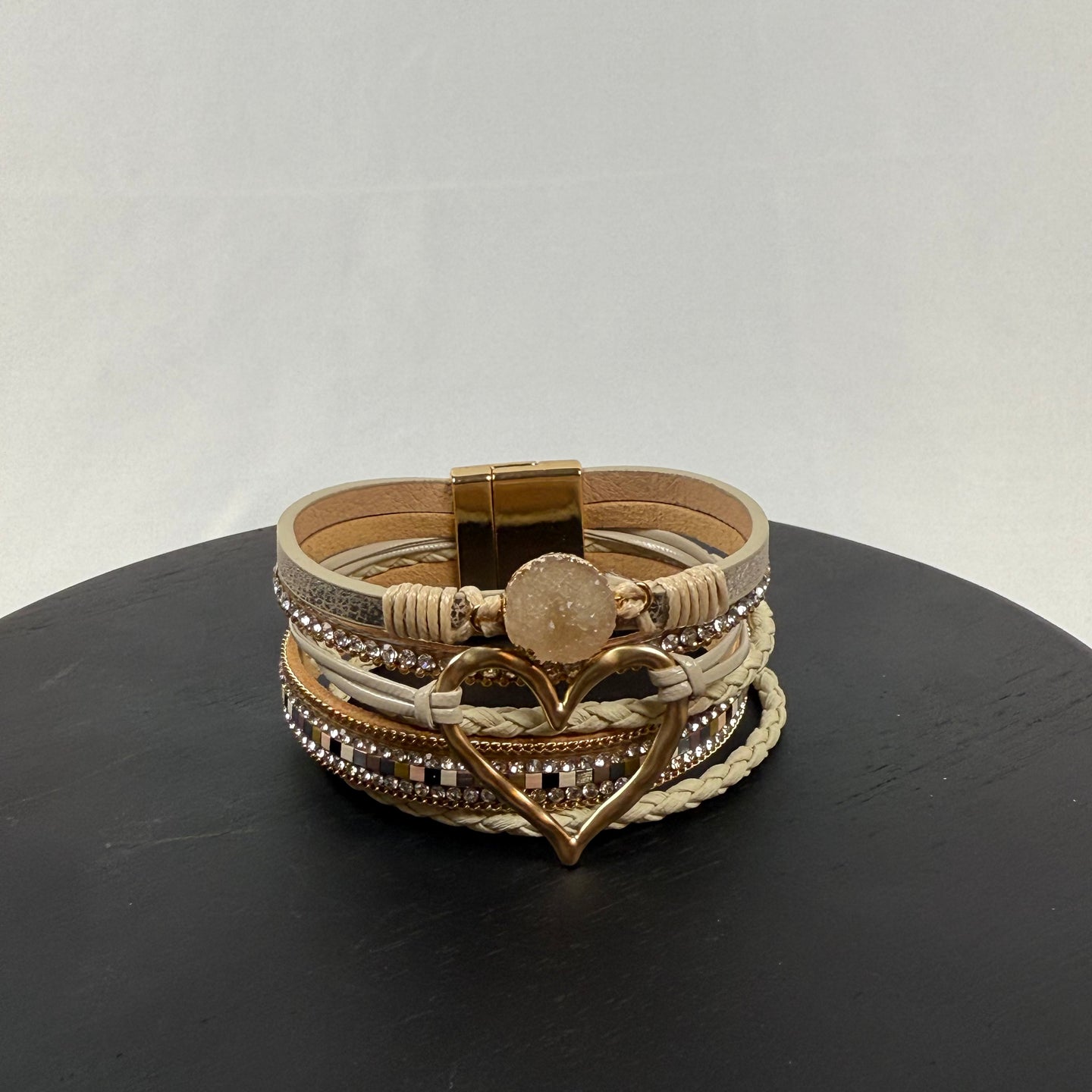 A beige and gold multi-layered bracelet with sparkling accents, braided sections, a round stone centerpiece, and a large heart-shaped charm, displayed on a black surface against a light background.