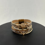 A beige and gold multi-layered bracelet with sparkling accents, braided sections, a round stone centerpiece, and a large heart-shaped charm, displayed on a black surface against a light background.