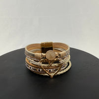 A beige and gold multi-layered bracelet with sparkling accents, braided sections, a round stone centerpiece, and a large heart-shaped charm, displayed on a black surface against a light background.
