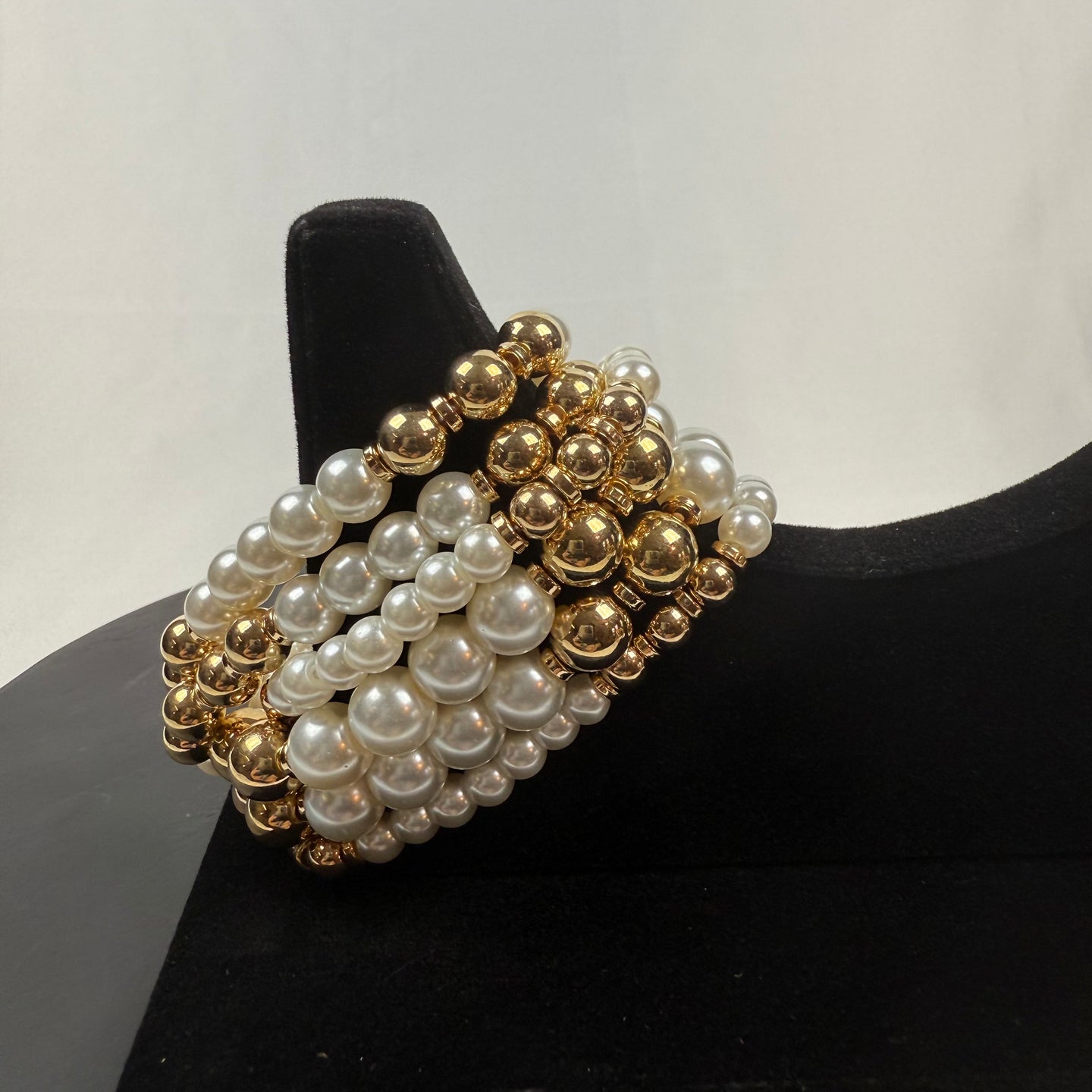 A bracelet made of alternating rows of shiny gold and white pearl-like beads displayed on a black velvet jewelry stand with a plain white background.