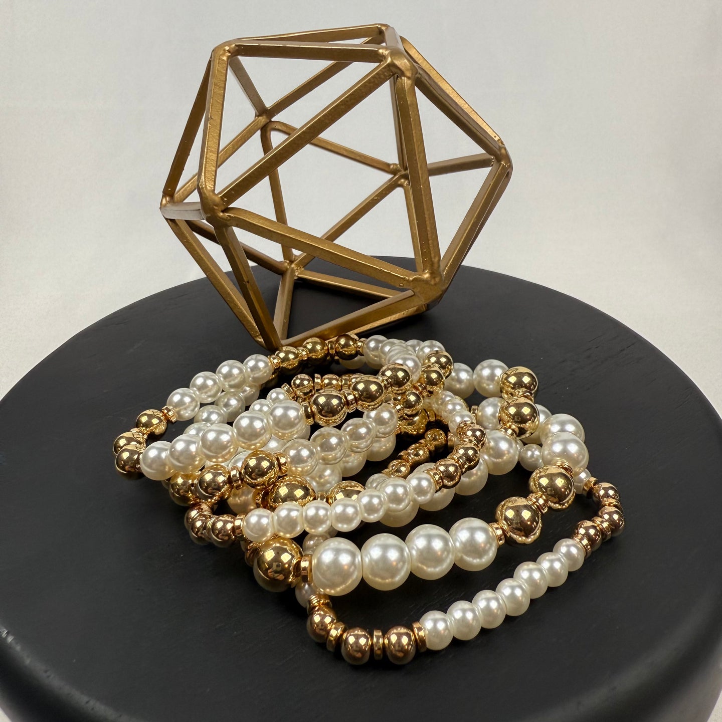 A geometric gold metal sculpture sits on a black circular base with several pearl and gold bead bracelets laid in front of it. The background is light and uncluttered.