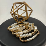 A geometric gold metal sculpture sits on a black circular base with several pearl and gold bead bracelets laid in front of it. The background is light and uncluttered.