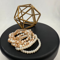 /Beige/ A geometric gold metal sculpture sits on a round black surface next to several strands of beige, white, and gold beads. The background is plain and white.