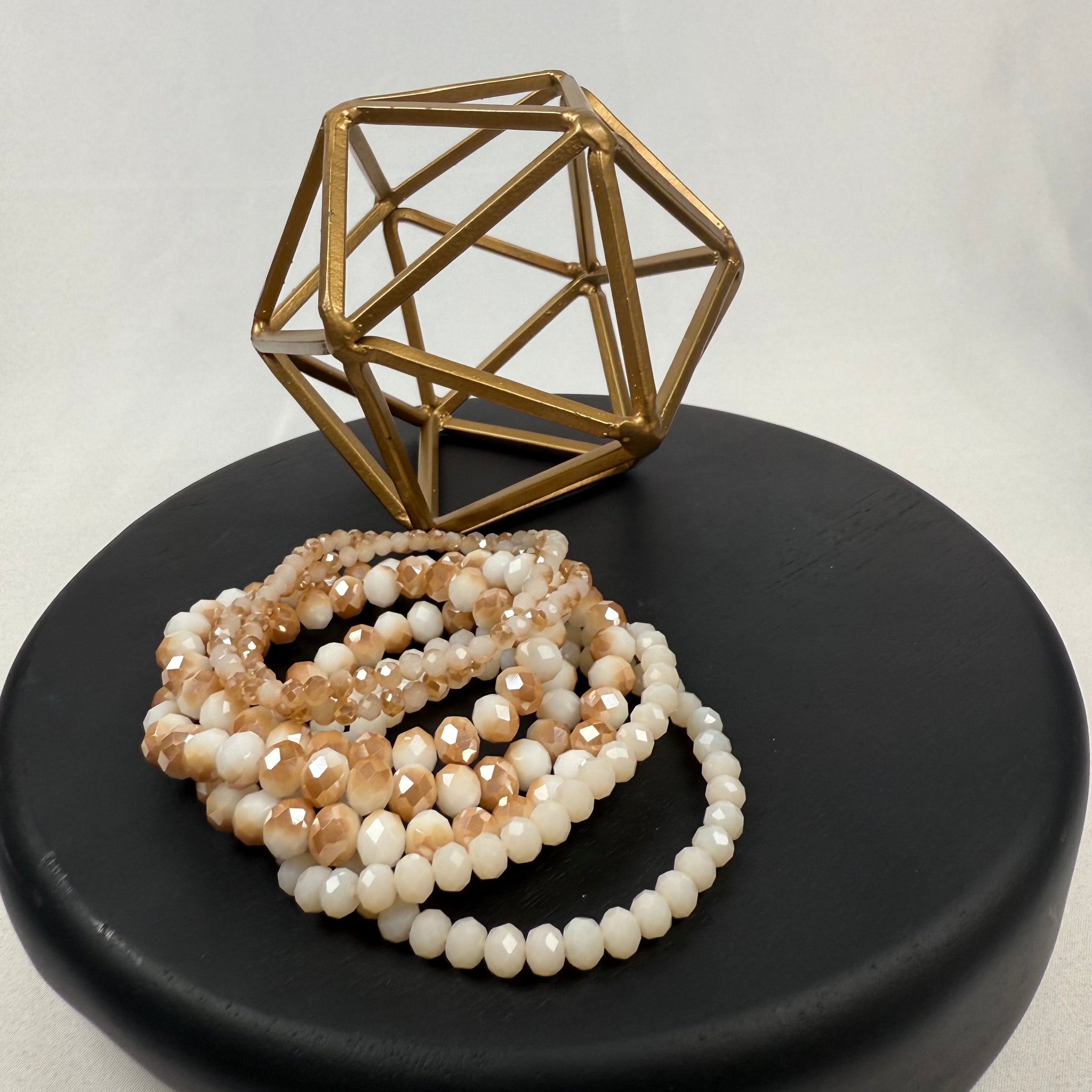/Beige/ A geometric gold metal sculpture sits on a round black surface next to several strands of beige, white, and gold beads. The background is plain and white.
