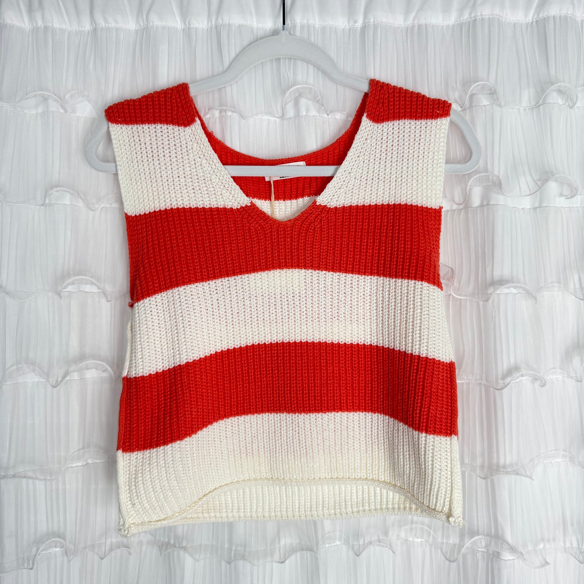 A sleeveless knit top with wide horizontal red and white stripes hangs on a clear hanger against a white, ruffled background. The top has a V-neck and a slightly cropped, boxy fit.