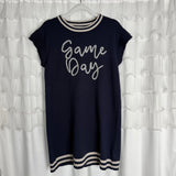 A navy blue short-sleeve dress with white trim on the collar, sleeves, and hem, featuring the words Game Day written in cursive on the front, hanging on a white hanger against a ruffled white backdrop.