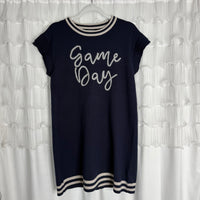 A navy blue short-sleeve dress with white trim on the collar, sleeves, and hem, featuring the words Game Day written in cursive on the front, hanging on a white hanger against a ruffled white backdrop.