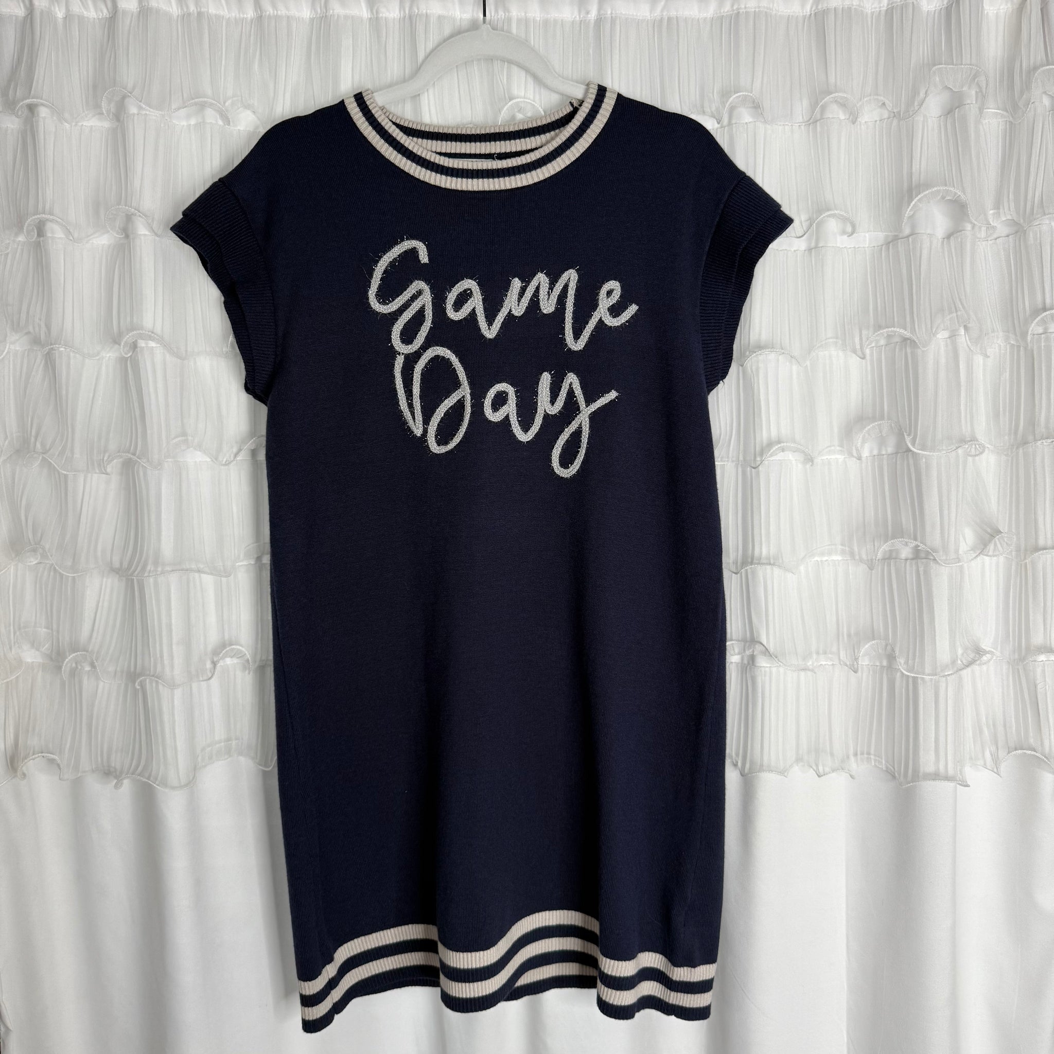 A navy blue short-sleeve dress with white trim on the collar, sleeves, and hem, featuring the words Game Day written in cursive on the front, hanging on a white hanger against a ruffled white backdrop.
