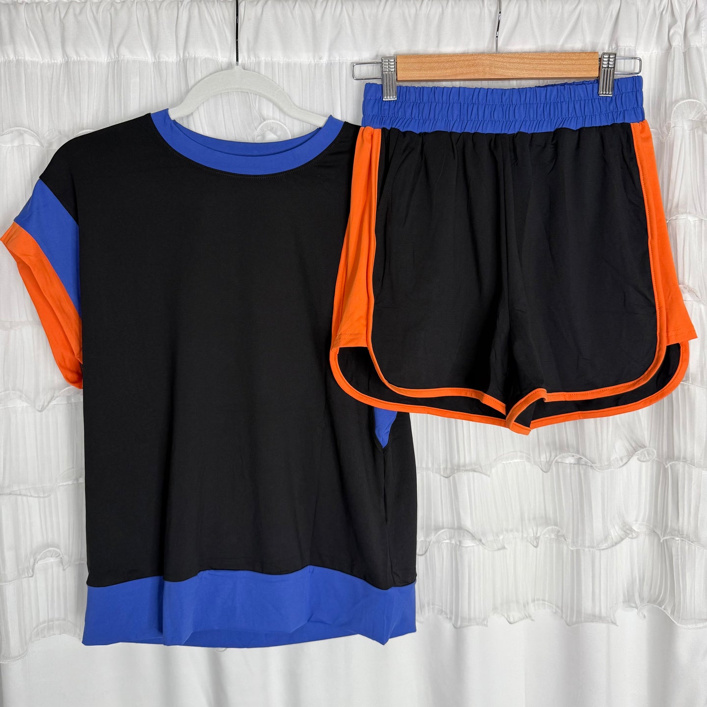 A matching athletic outfit hangs on hangers: a short-sleeve black shirt with blue accents and a pair of black shorts bordered with bright orange and blue trim, both displayed against a white ruffled curtain.