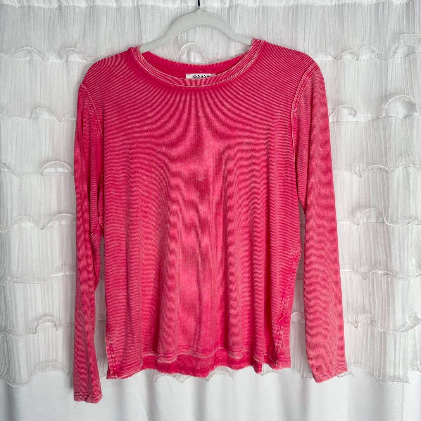 /Fuschia/ A long-sleeve, faded pink shirt hangs on a clear plastic hanger in front of a white textured curtain. The shirt has a ribbed neckline and visible stitching along the hems.