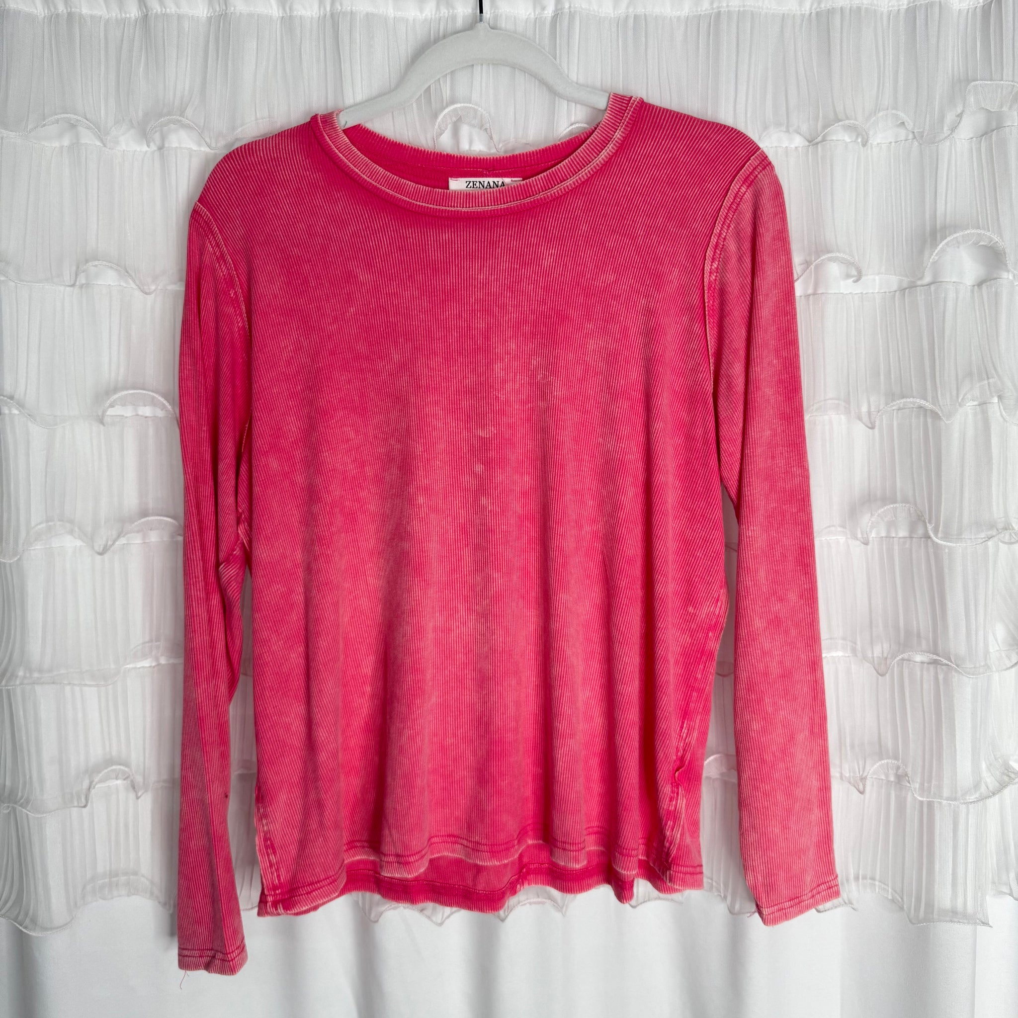 /Fuschia/ A long-sleeve, faded pink shirt hangs on a clear plastic hanger in front of a white textured curtain. The shirt has a ribbed neckline and visible stitching along the hems.