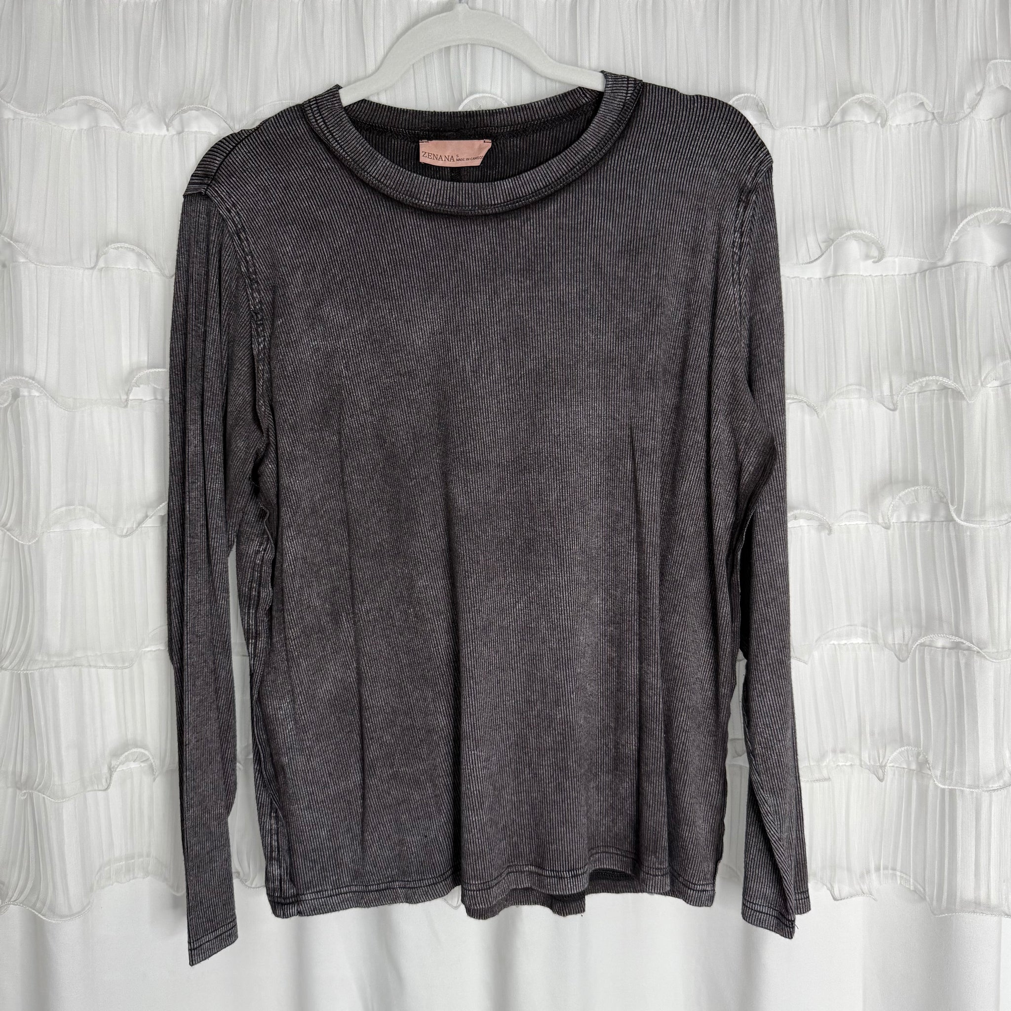 /Ash Black/ A long-sleeved, dark gray ribbed shirt hangs on a clear hanger in front of a white, textured curtain background.