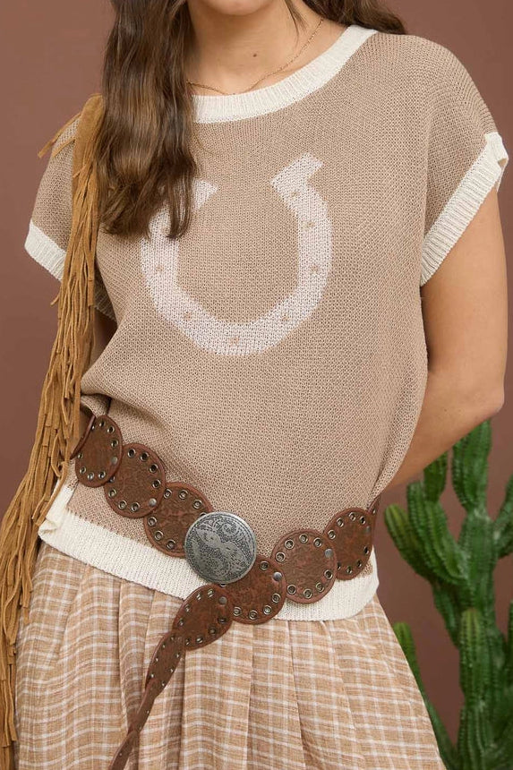 Woman wearing a beige knit top with a horse shoe design, plaid skirt, and brown belt against a brown background.