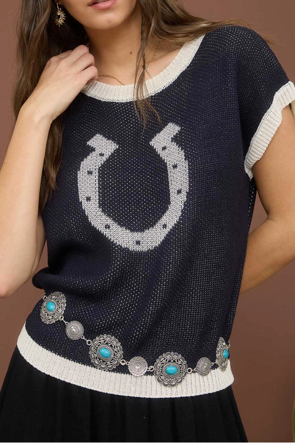 Woman wearing a navy blue sweater with a horseshoe design and a black hat with turquoise accents on a brown background.