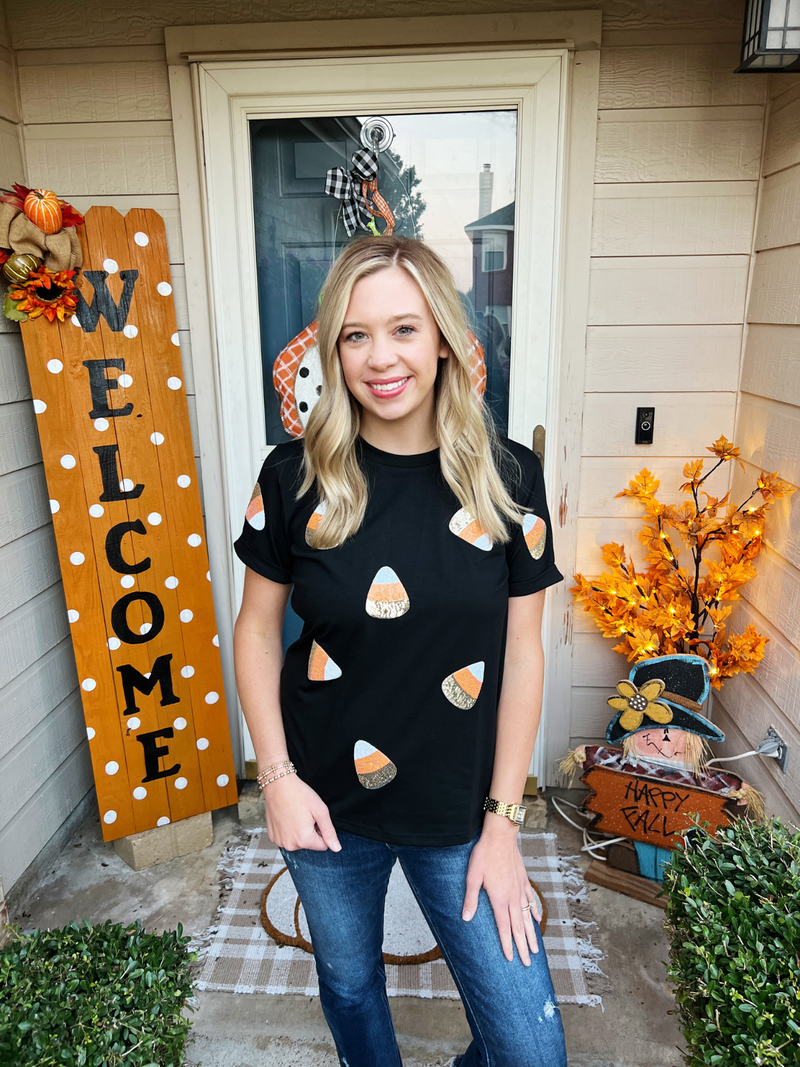 Woman wearing a black t-shirt with candy corn pattern standing in front of a 'Welcome' sign and autumn decorations.